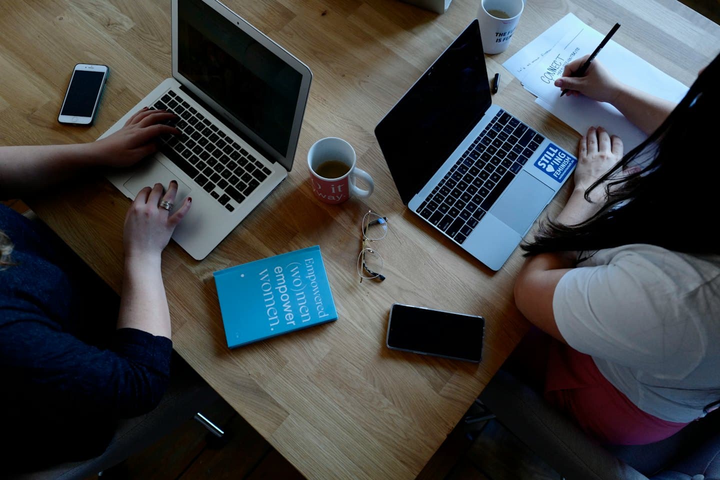 Two people collaborate at a wooden table with laptops and notebooks.